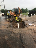Backhoe and crew working at an excavation in a paved lot.