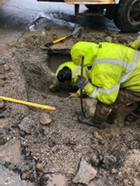 Crew member in high-visibility rain gear digging by hand in a muddy excavation.