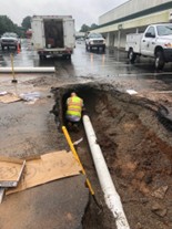 Crew member in a high-visibility vest working in a long trench beside a white pipe on a wet street.