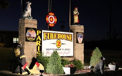 Lighted sign reading Firehouse 2 and September 11, 2001, with firefighter statues, at night.