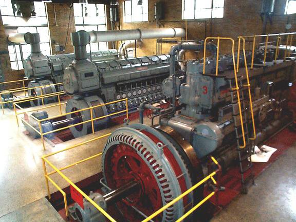 Row of large diesel generator engines with red generator housings and yellow safety railings inside the power plant.