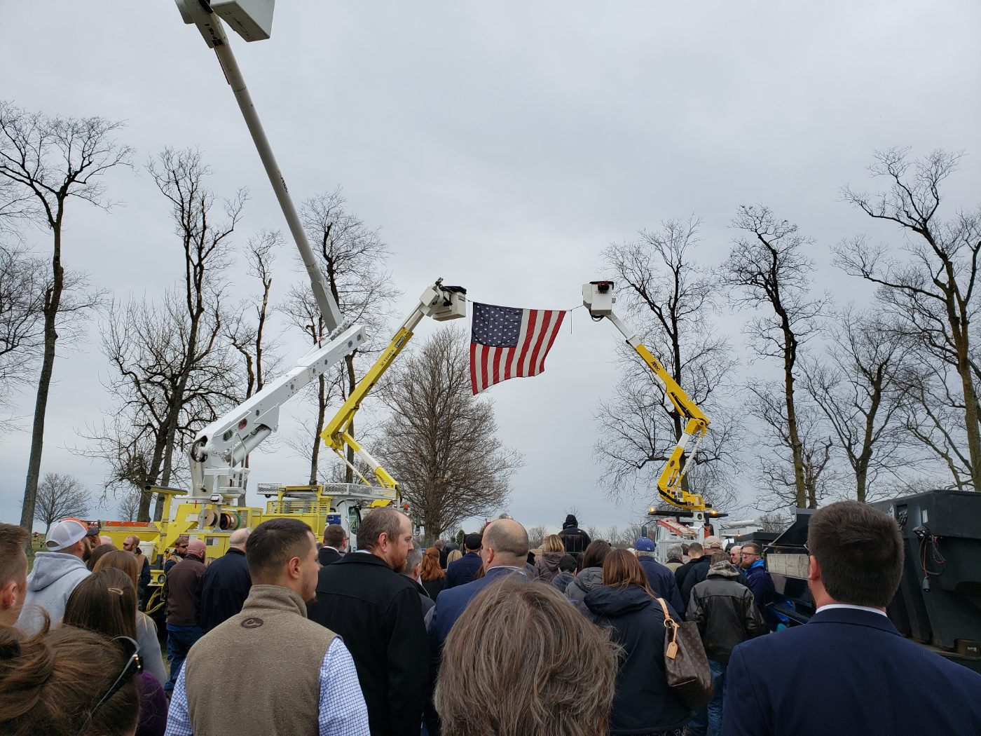 An American flag hung between two raised bucket trucks above a gathering of people.
