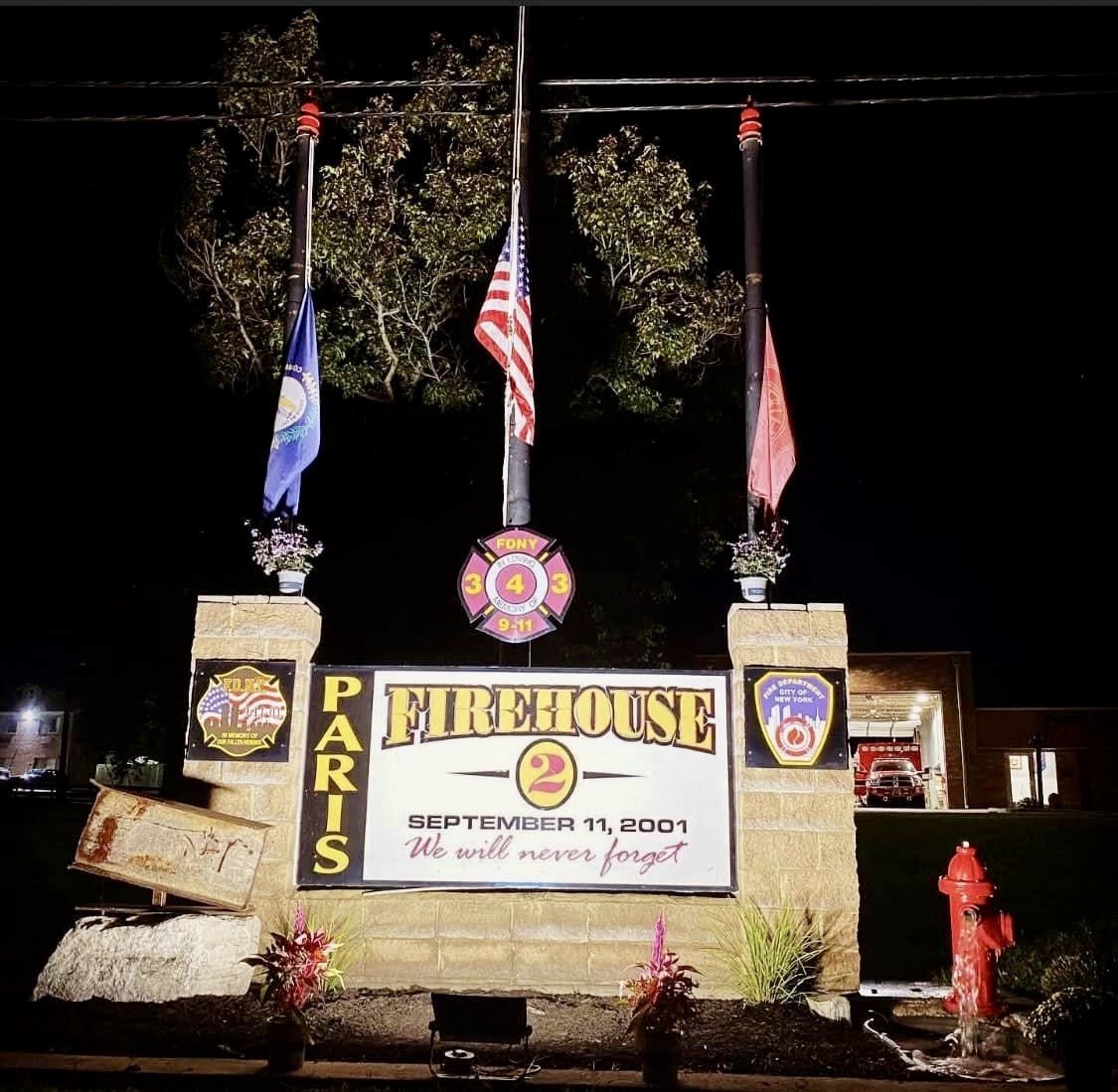 Fire Station 2 sign at night reading Firehouse 2, September 11, 2001, We will never forget, with flags and a red fire hydrant.