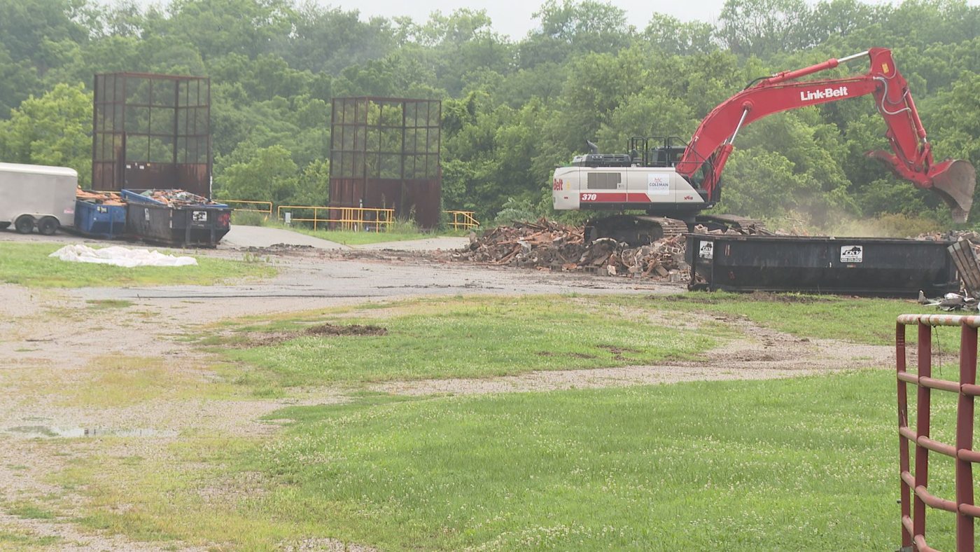 Excavator tearing down the former transfer station, with piles of brick rubble, a roll-off dumpster, and the tall caged loading structures in the background.